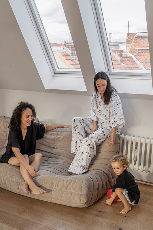 dos mujeres sentadas en el sofa, una con un conjunto negro Jinbei en viscosa de Forever 2 y otra con blusa y pantalón estampados de Forever 2 en algodón, y niña jugando con un globo rojo